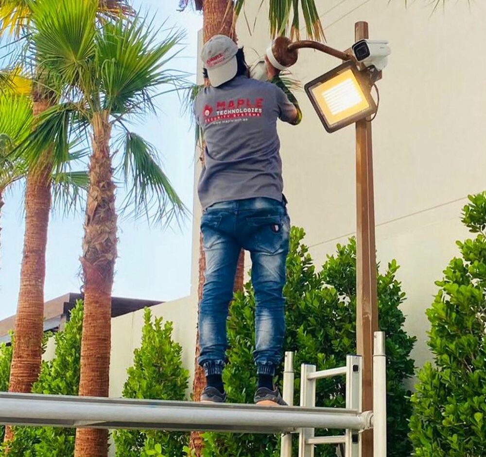 A MapleTech technician installing a camera and floodlight on a pole among palm trees